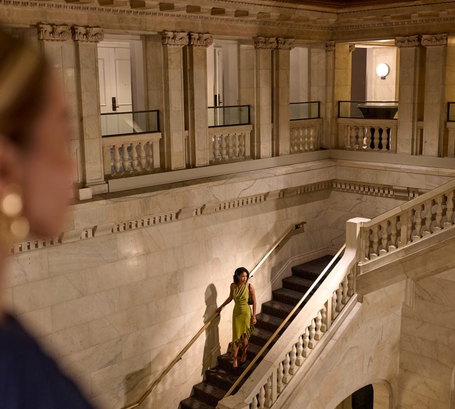 Woman in a lime green dress descends a marble staircase in an elegant, classical interior with ornate columns and railings.