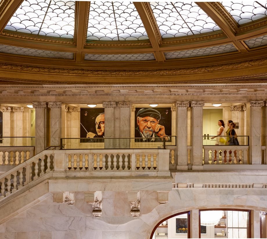 Grand marble interior featuring a sweeping staircase, ornate balustrades, and large portrait paintings beneath a decorative skylight ceiling.