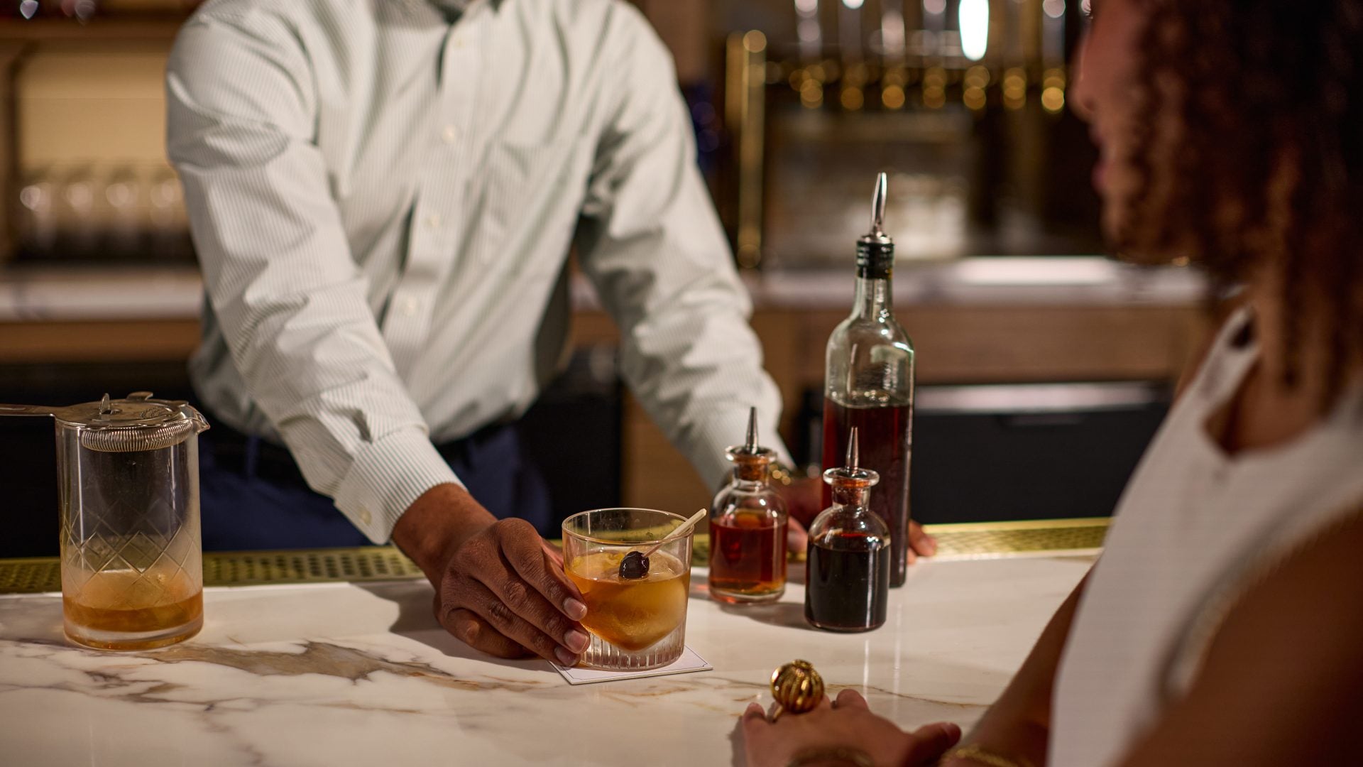 Bartender hands an orange cocktail to a customer at a marble bar, with liquor bottles in the background.