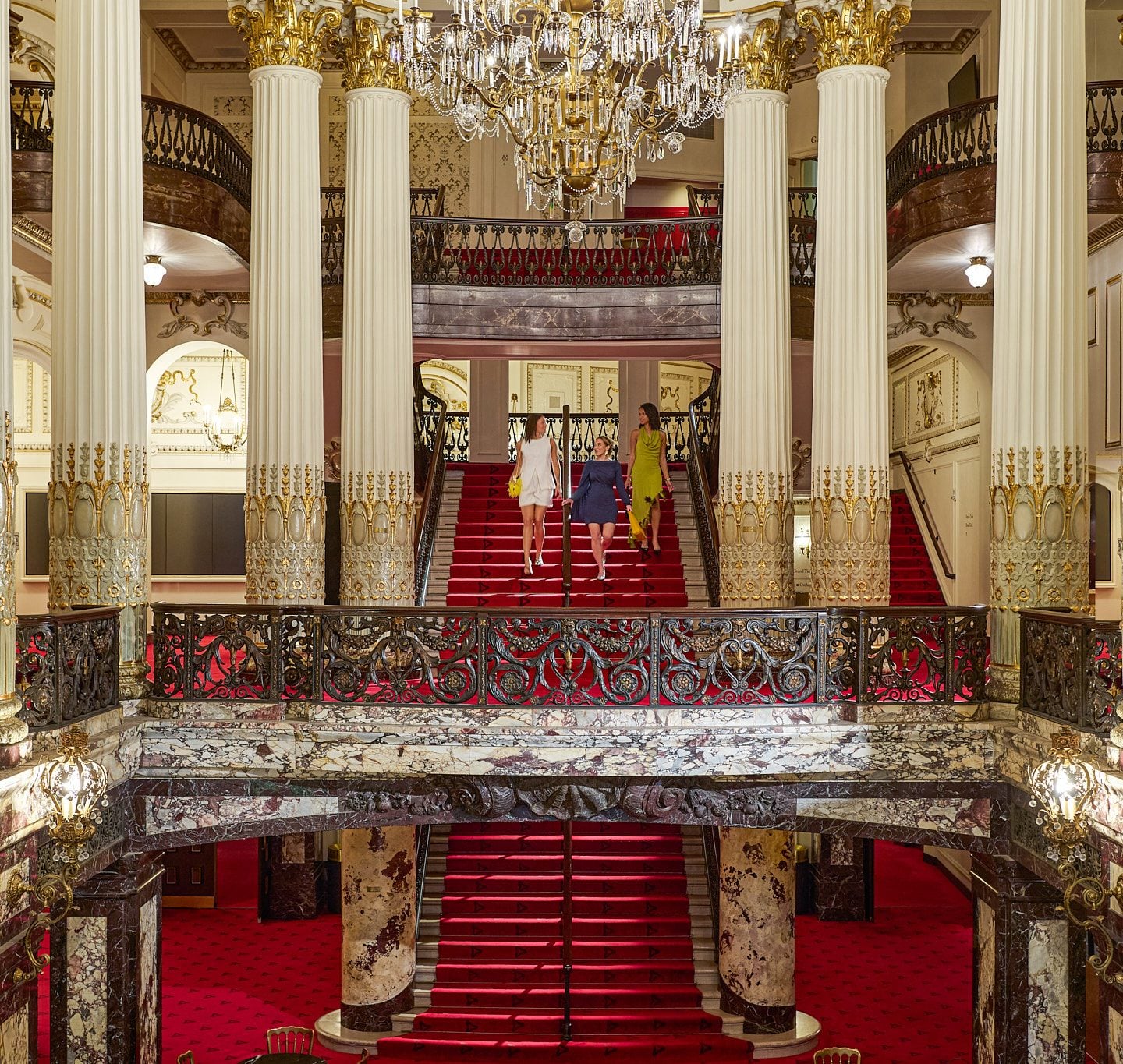 Grand opulent lobby with a red carpet staircase and crystal chandeliers; three people descend the stairs in stylish dresses.