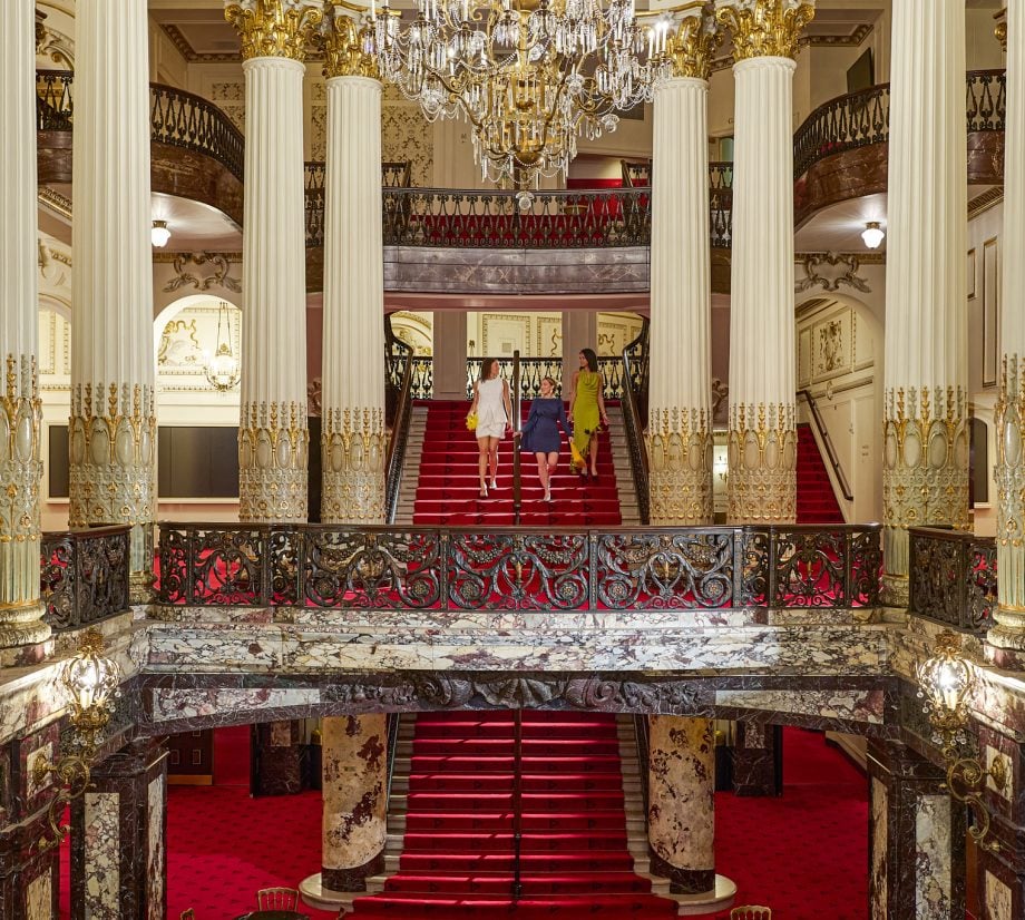Grand opulent lobby with a red carpet staircase and crystal chandeliers; three people descend the stairs in stylish dresses.