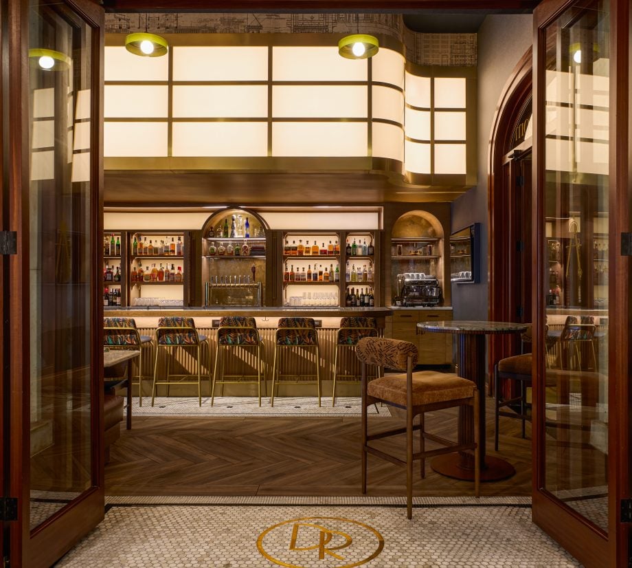 Open glass-door entrance to a warm, upscale bar with a long liquor display and gold bar stools inside, logo on tiled floor.