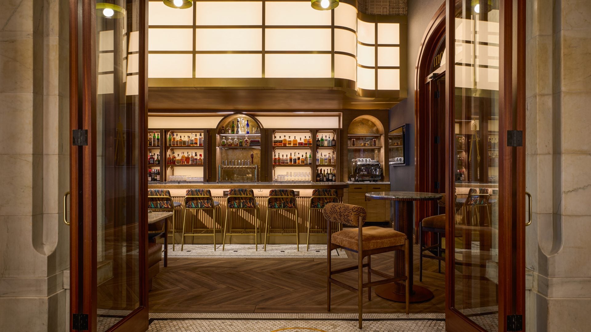 View of a warmly lit hotel bar through open glass doors, with backlit liquor shelves and brass stools along the counter.