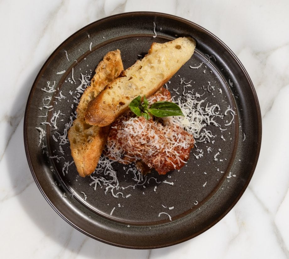 Plate of pasta with tomato sauce, grated cheese, and crusty bread slices, garnished with basil on a dark plate over a marble surface.