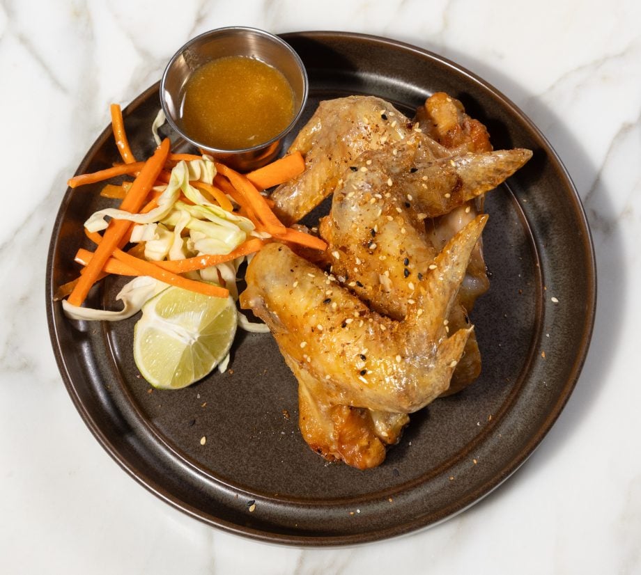 Plate with sesame-seasoned roasted chicken, carrot and cabbage slaw, lime wedge, and a small cup of dipping sauce on a dark plate.
