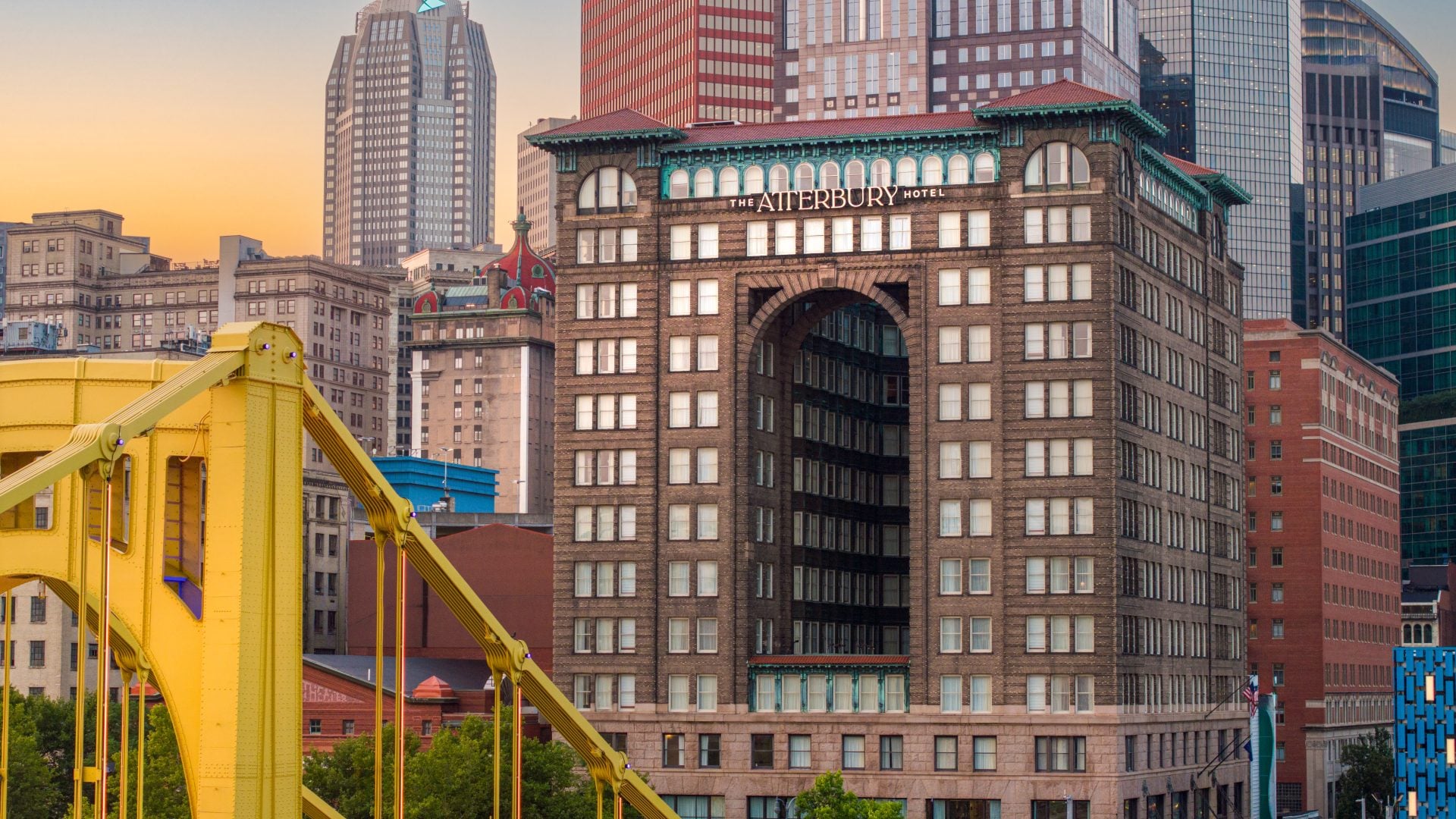 Sunset cityscape featuring The Atterbury Hotel’s arched gateway and a yellow bridge in the foreground among modern skyscrapers.