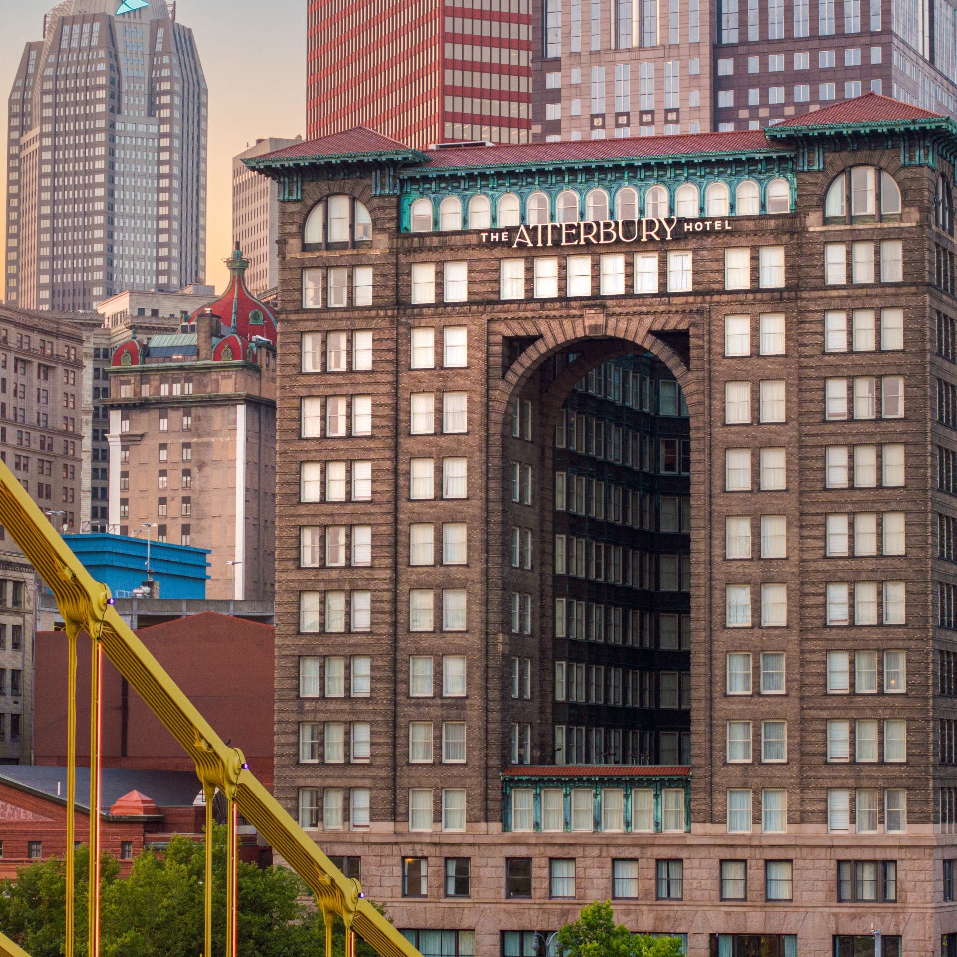 Sunset cityscape featuring The Atterbury Hotel’s arched gateway and a yellow bridge in the foreground among modern skyscrapers.