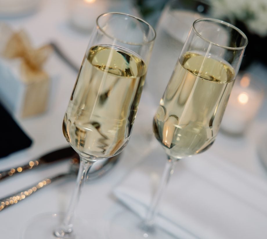 Two champagne flutes filled with bubbly, raised in a toast on a white-tabletop banquet setting with blurred flowers in the background, suggesting a celebration.