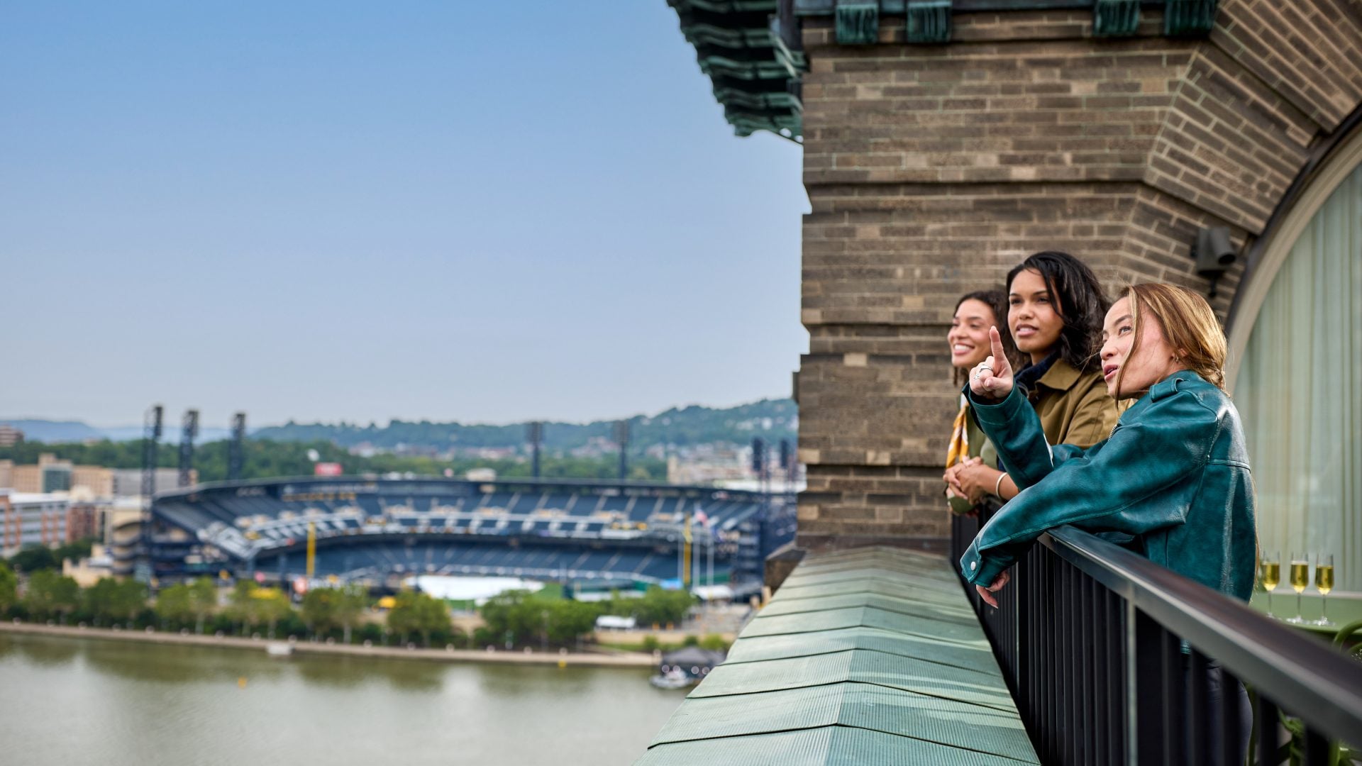 Three women stand at a balcony rail on a brick building, pointing toward a stadium and city skyline in the distance by a river.