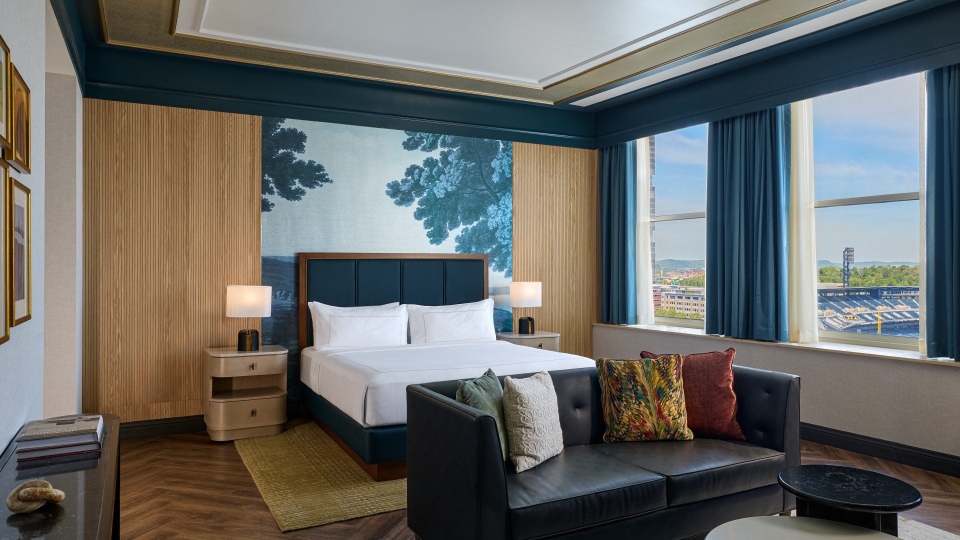 Hotel bedroom with a white bed, teal headboard, wood-paneled walls, and a seating area by large windows with blue curtains.