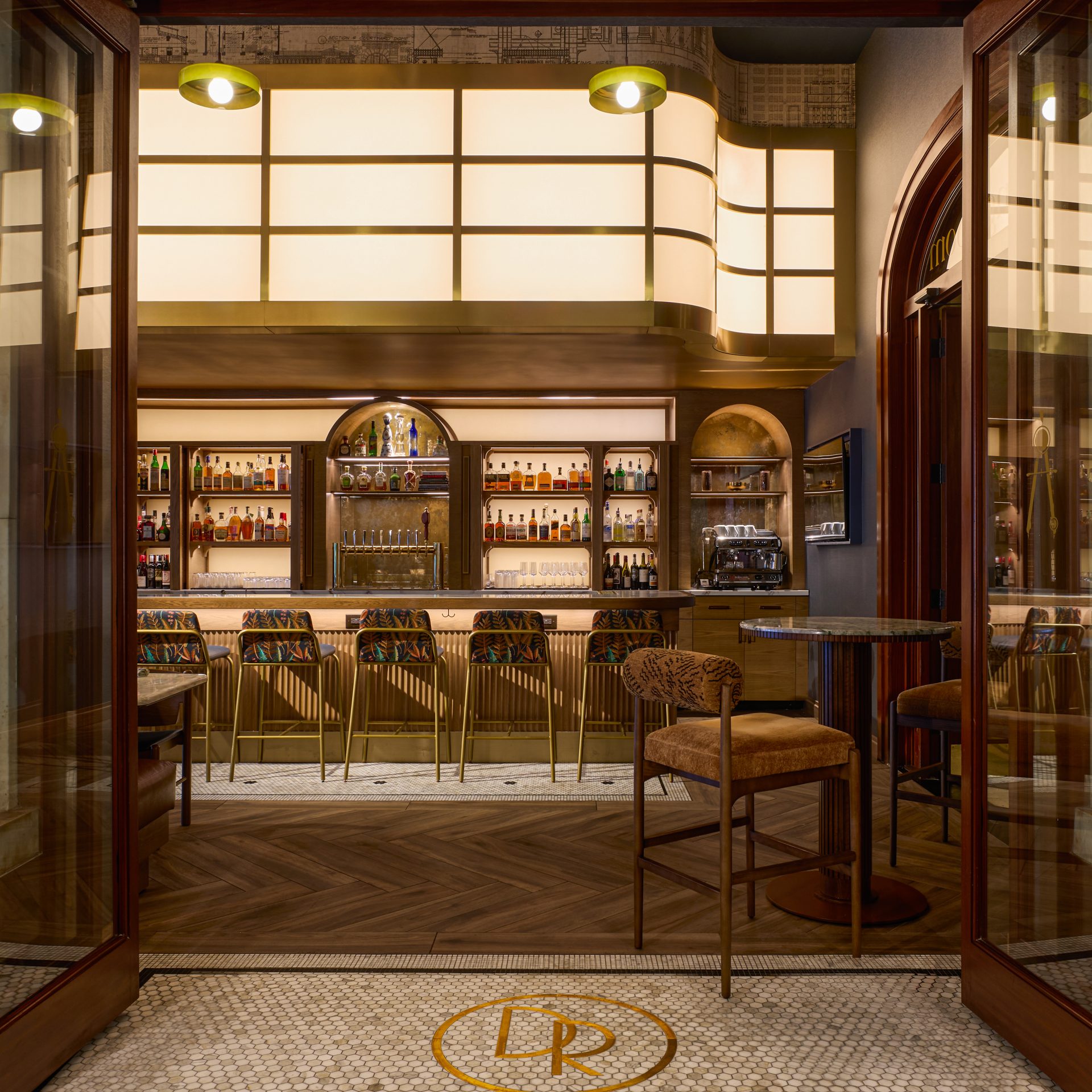 Open glass doors frame a classy hotel bar with backlit shelves stocked with bottles and tall stools along the counter at center.
