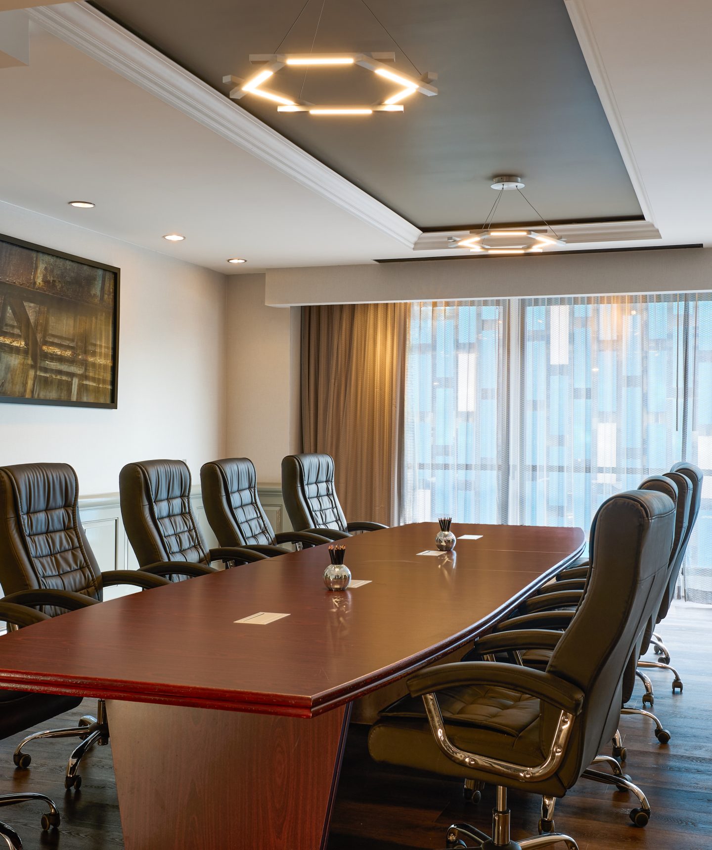 Boardroom with a long wooden table surrounded by black leather high-back chairs, large windows with sheer curtains behind it.