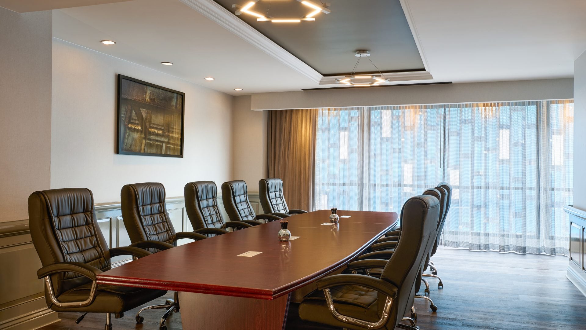 Boardroom with a long wooden table surrounded by black leather high-back chairs, large windows with sheer curtains behind it.
