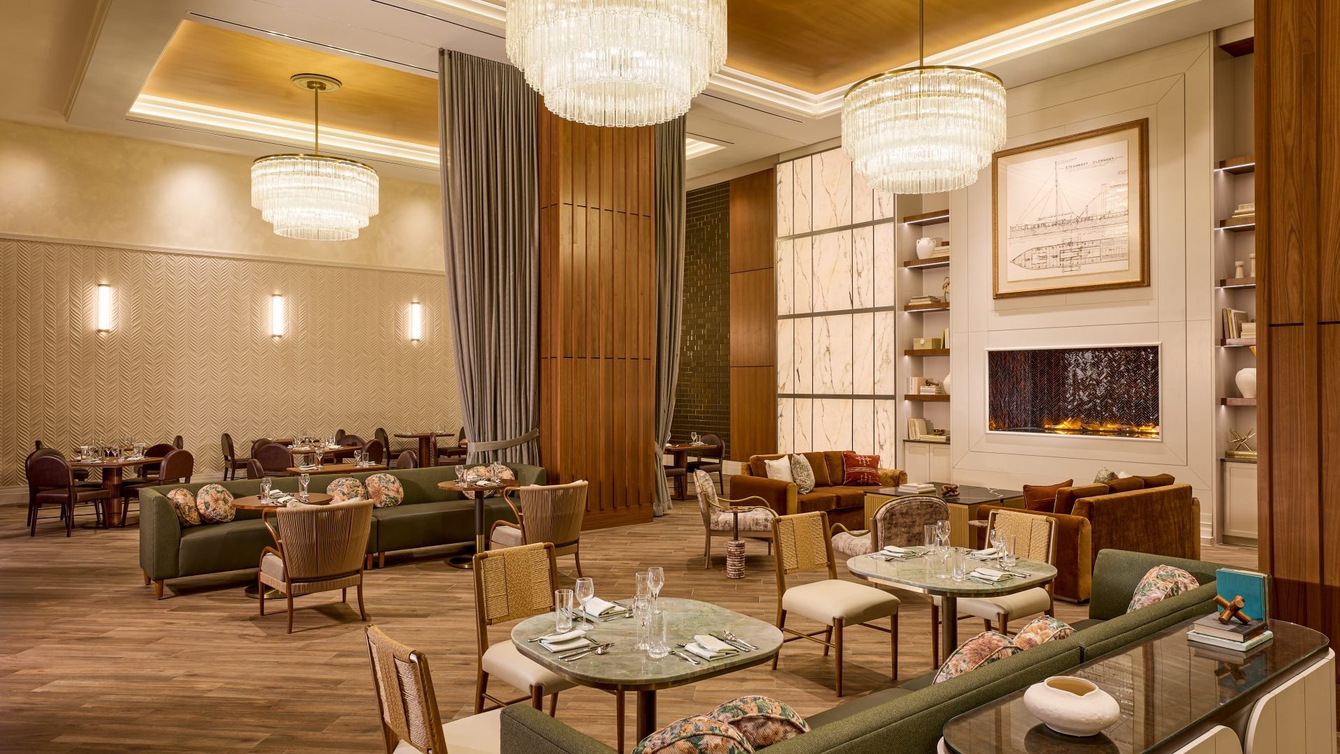 Elegant hotel restaurant lounge with large crystal chandeliers, wooden columns, and plush green sofas around marble tables in warm lighting.