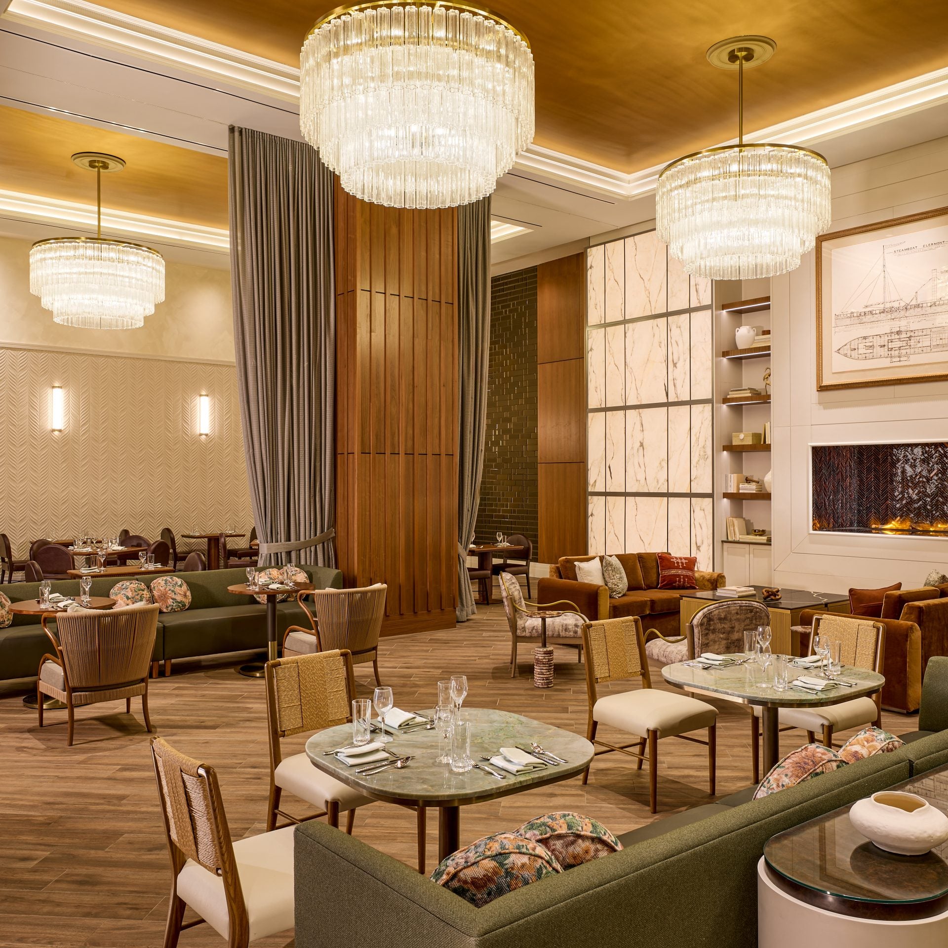 Elegant hotel restaurant lounge with large crystal chandeliers, wooden columns, and plush green sofas around marble tables in warm lighting.