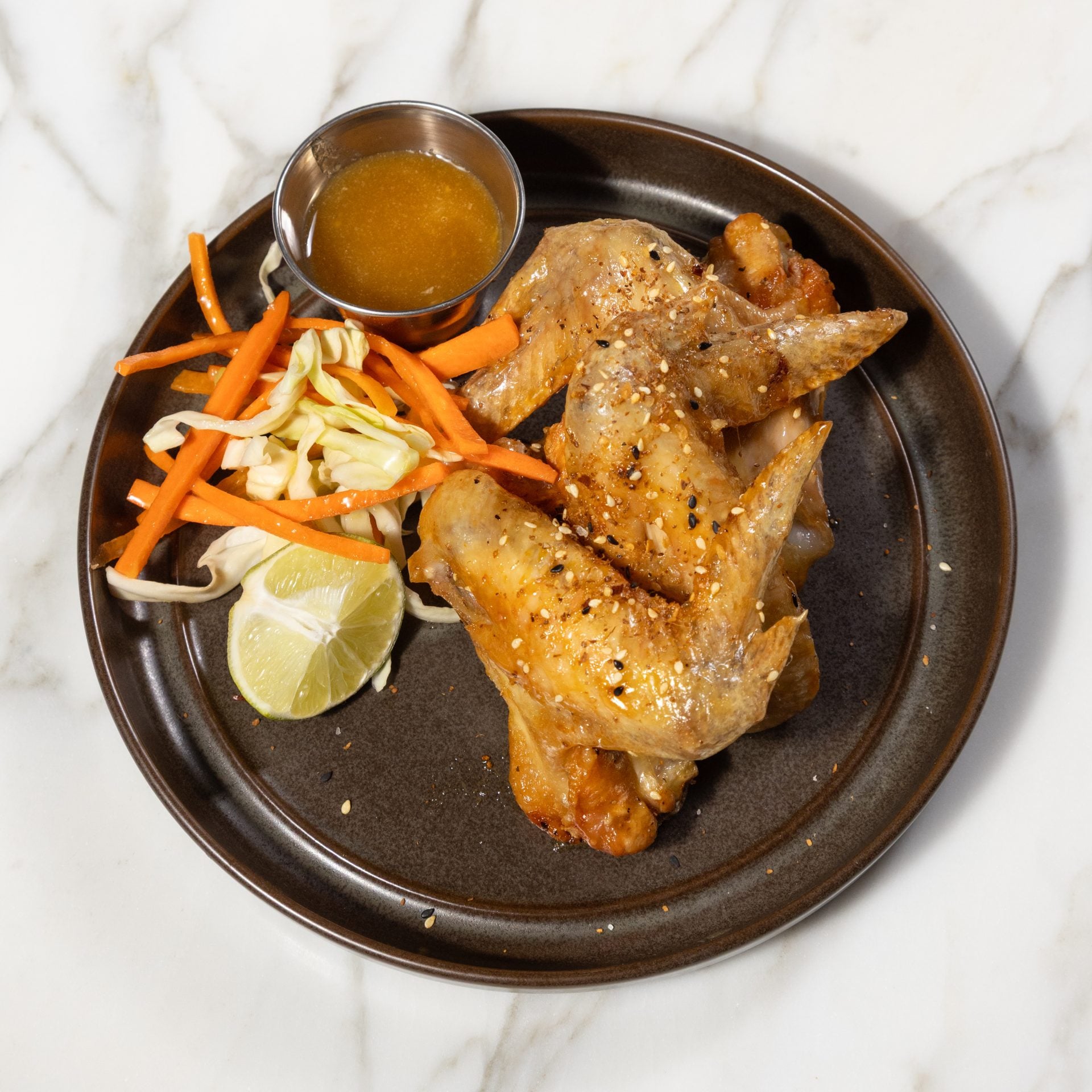 Plate with glazed chicken wings sprinkled with sesame seeds, served with carrot-jullien slaw, lime wedges, and sauce in a metal cup on a dark plate.
