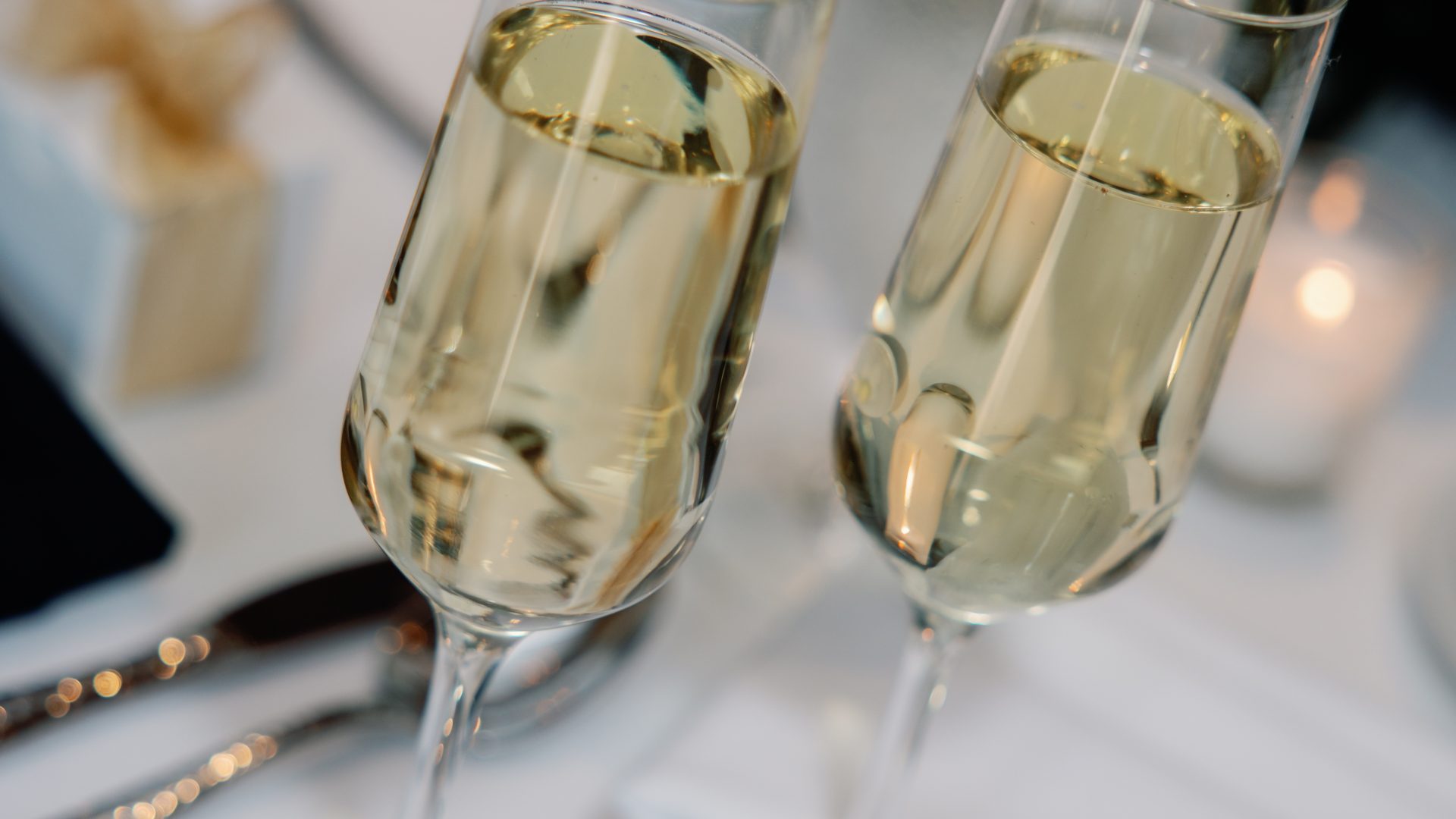 Two champagne flutes filled with sparkling wine tilted together for a toast on a white-draped table with blurred floral decor and candlelight in the background.