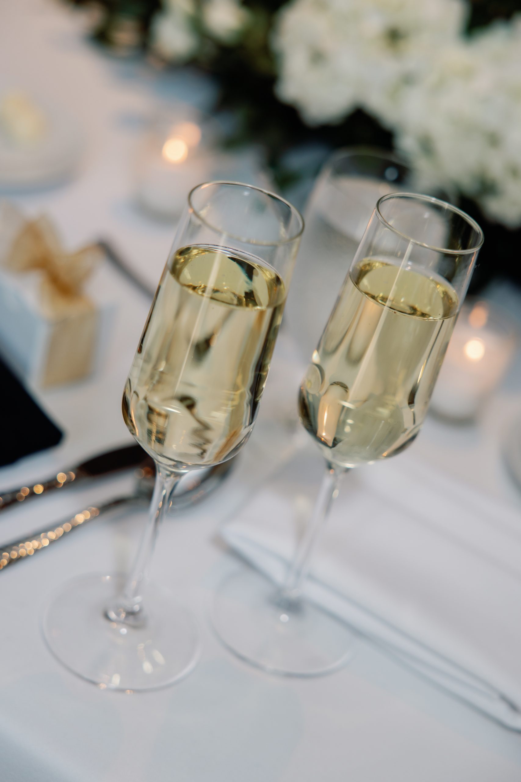 Two champagne flutes filled with sparkling wine tilted together for a toast on a white-draped table with blurred floral decor and candlelight in the background.